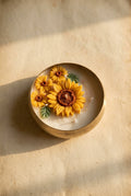 A hand holds an open gift box containing a round white candle decorated with three yellow sunflowers, nestled in brown crinkle paper packaging.