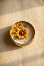A hand holds an open gift box containing a round white candle decorated with three yellow sunflowers, nestled in brown crinkle paper packaging.