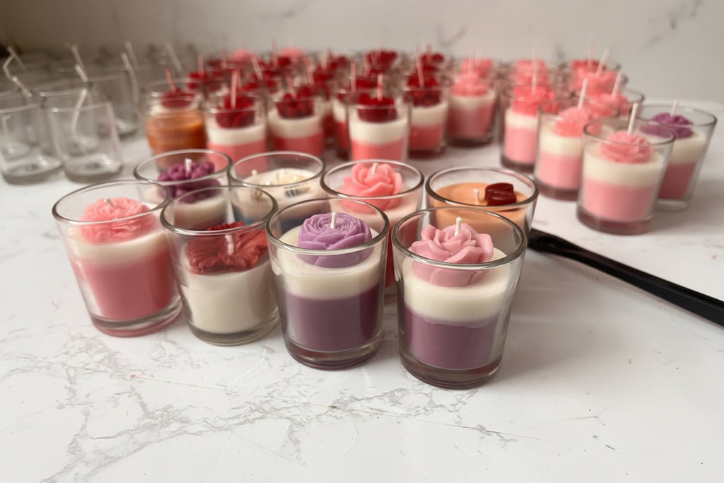 A collection of small glass jar candles arranged on a light marble-like surface. The candles feature layered wax designs, including pink and white, purple and white, red and white, and peach and white, with some topped by red rose-shaped wax ornaments.