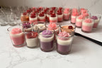 A collection of small glass jar candles arranged on a light marble-like surface. The candles feature layered wax designs, including pink and white, purple and white, red and white, and peach and white, with some topped by red rose-shaped wax ornaments.
