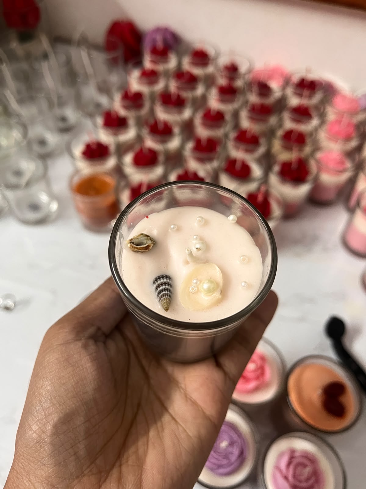 A hand holds a small glass jar candle with a pale pink wax body and decorative shell and pearl accents. The background shows many similar candles arranged on a table, with some red flower toppers visible.