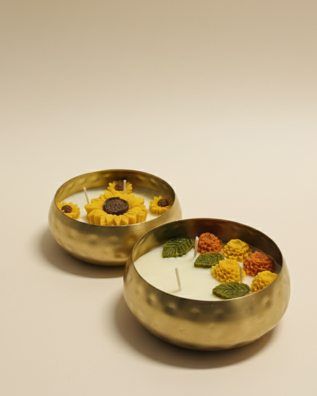 A high-angle, professional studio shot of two luxury candles in round hammered gold brass bowls. The foreground candle features cream-colored wax topped with intricate hand-sculpted orange and yellow wax flowers with green leaves. The background candle features large yellow wax sunflowers. The background is a clean, neutral cream tone with soft, elegant lighting.