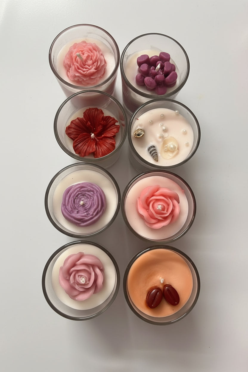 Eight decorative soy candles in clear glass jars arranged on a white marble surface. The candles feature various floral and textured wax tops in pink, red, purple, peach, and dark red, with one candle showing a pearl and shell detail.