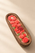 Several elongated wooden trays containing colorful decorative items, including pink and coral flowers, green leaves, shells, and small pearl-like beads, arranged on top of newspaper.
