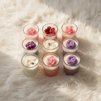 A collection of colorful layered wax candles in clear glass jars, arranged in rows on a white marble surface. The candles feature various color combinations including pink, red, white, purple, and blue, with some decorated with small flower-shaped wax tops.