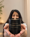 A woman holds a lit black candle jar labeled 'LUMINAURA WOODY VANILLA Bergamot • Sandalwood' toward the camera in an indoor setting.