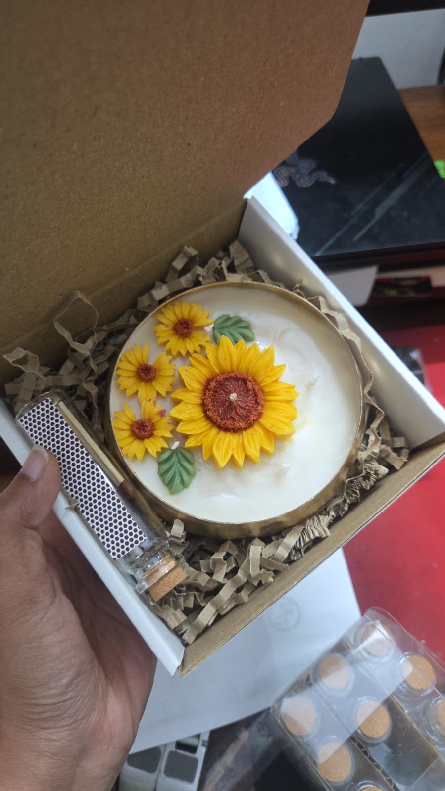 A hand holds an open gift box containing a round white candle decorated with three yellow sunflowers, nestled in brown crinkle paper packaging.