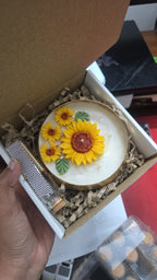 A hand holds an open gift box containing a round white candle decorated with three yellow sunflowers, nestled in brown crinkle paper packaging.