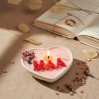 Heart-shaped pink candle with red letters spelling 'MAA' on a white surface with decorative elements.